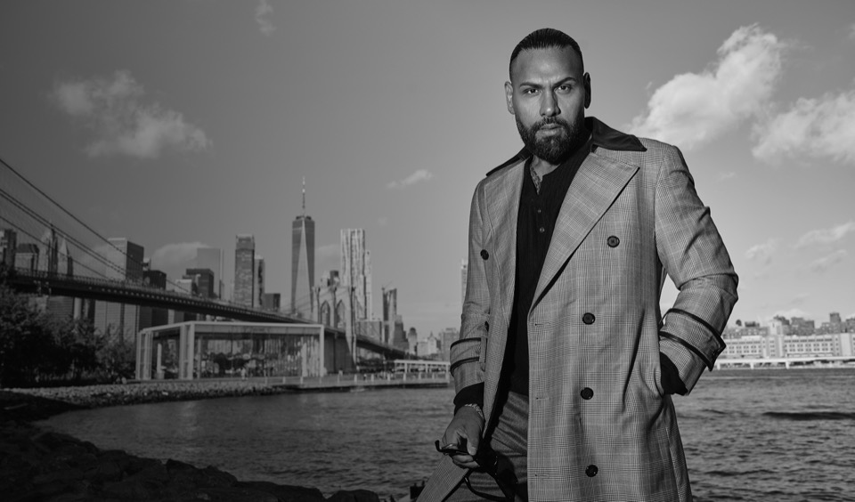 Jas Mathur in a tailored overcoat on the Brooklyn waterfront, with Manhattan behind him